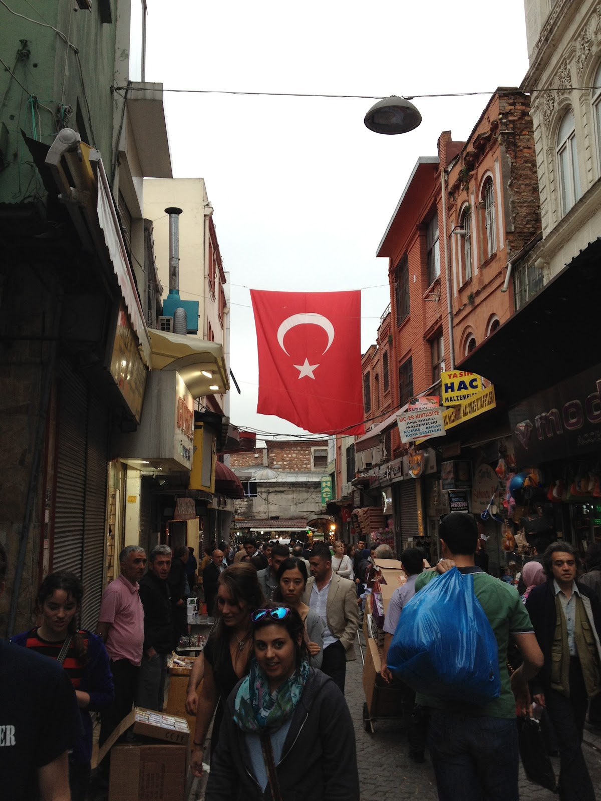 Market place, people are buying and walking in the market and a Turkey flag can be seen. 