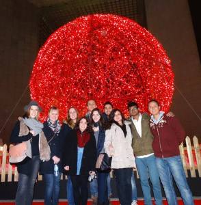 A group of 10 people standing Infront of a huge red decorated ball during night. 